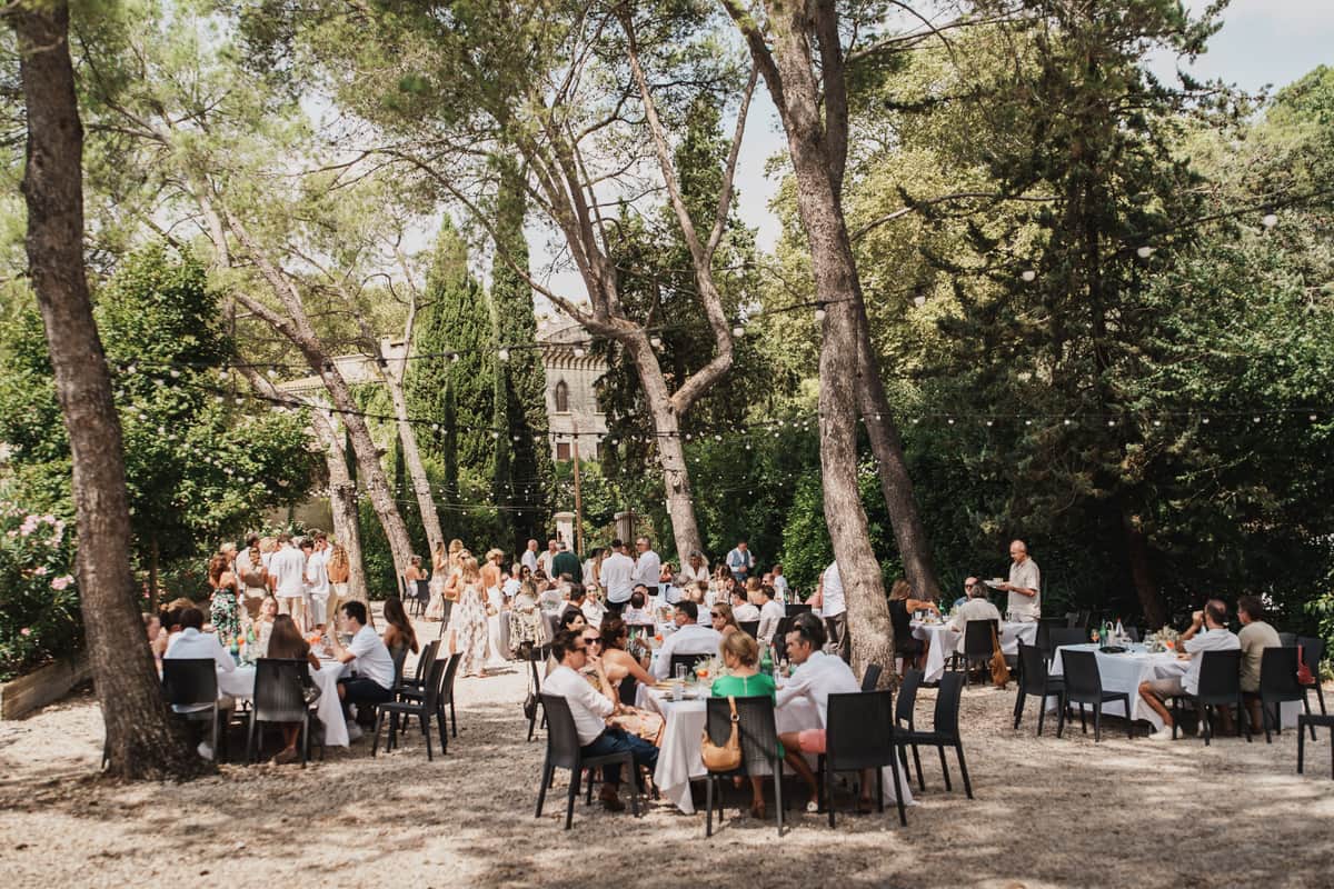 Guests dining at white-clothed tables under tall pine trees strung with festoon lights, a stone building beyond.