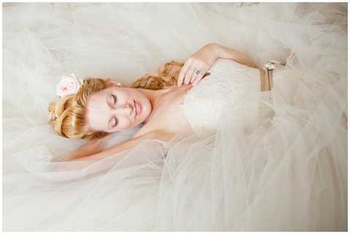 A bridal portrait shot from above showing the bride lying down surrounded by the voluminous tulle skirt of her strapless ivory lace gown, which fans out to fill nearly the entire frame. The bride has wavy auburn hair adorned with a blush pink flower hair accessory, her eyes are gently closed, and she wears a blue gemstone ring on her hand resting near her shoulder. The composition is an overhead close-up portrait with soft, bright, high-key lighting that gives the image a light and airy feel.