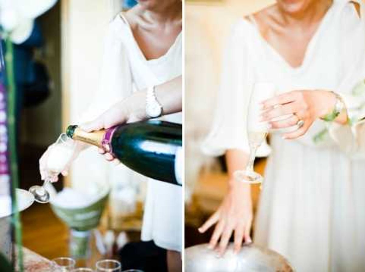 A diptych of two close-up shots capturing a woman in a white dress pouring champagne from a dark green bottle. In the left frame, she tilts the bottle over a champagne flute near what appears to be an ice bucket in a warmly lit interior setting. The right frame shows a tighter detail shot of her hand holding a filled champagne flute, with a large cocktail ring visible on her finger and a small floral accessory or boutonniere nearby. Both shots are styled as getting-ready or pre-ceremony moments, with a soft, bright editorial feel.