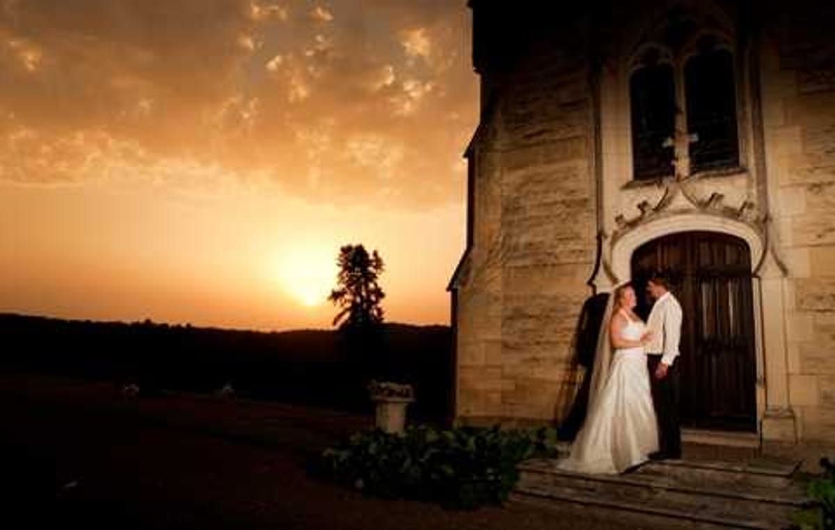 A couple poses together at the entrance of a stone chapel or chateau with Gothic-style arched doorway and decorative stonework, photographed at sunset with a vivid orange sky in the background. The bride wears a white floor-length gown and the groom is dressed in a light-colored suit, and they are leaning close together on the stone steps of the entrance. The wide-angle shot uses dramatic backlighting from the setting sun, creating strong contrast between the warm golden sky and the darkened silhouette of the building and surrounding landscape. Potential venue feature image.