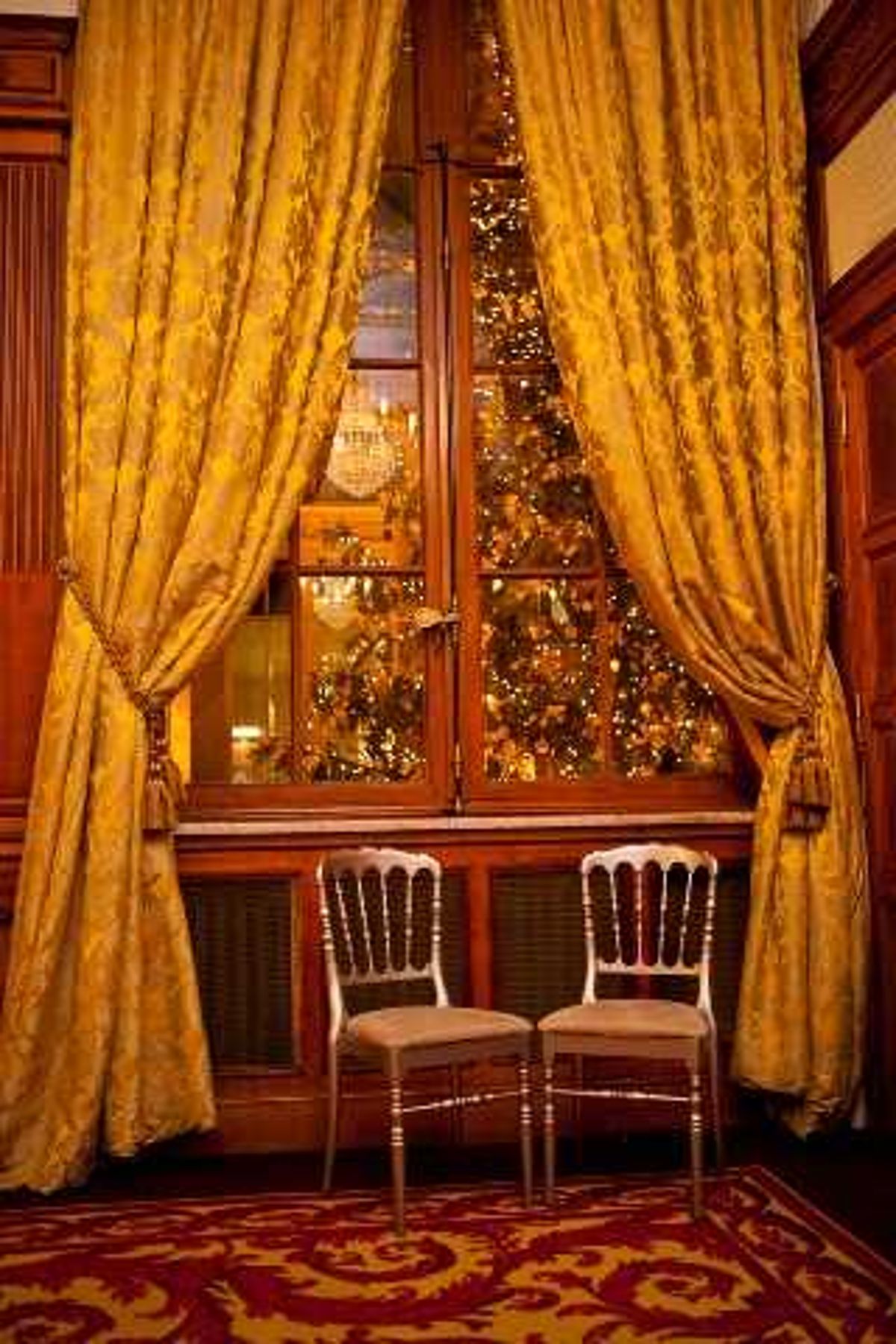 Detail shot of an interior venue space featuring two gold Chiavari chairs positioned side by side in front of a tall wooden-framed window flanked by floor-length gold damask curtains with tassel tiebacks. Through the window, a warmly lit Christmas tree decorated with golden fairy lights is visible, suggesting a winter or holiday-season wedding. The flooring is covered in a deep red and gold patterned carpet, and the wood-paneled walls contribute to a classic, formal interior aesthetic. The overall decor palette is rich gold and burgundy. Potential venue feature image.