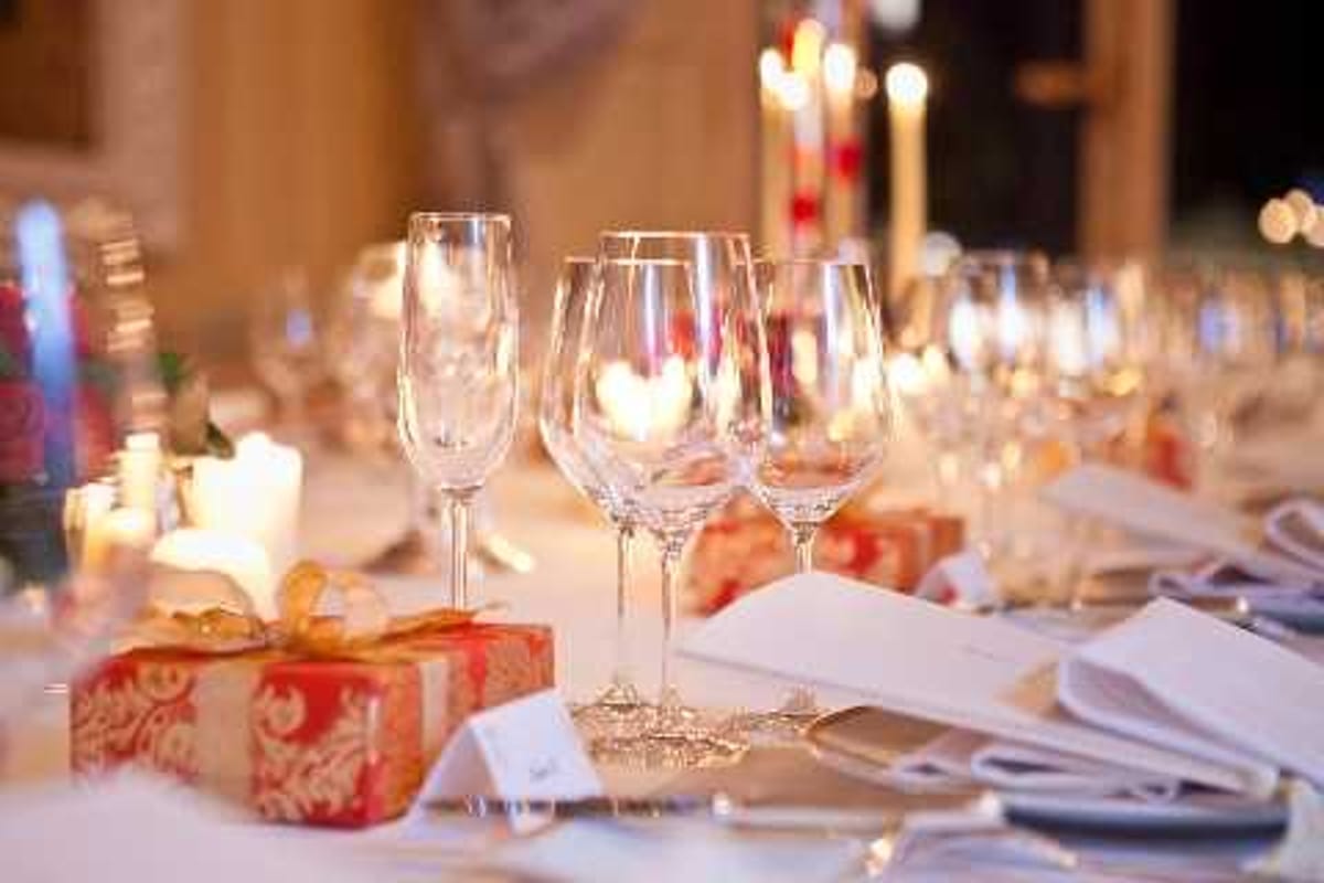 Close-up detail shot of a wedding reception table setting in a warmly lit indoor venue. The table is dressed in white linens with crystal champagne flutes and wine glasses arranged at each place setting, accompanied by folded white napkins and silver cutlery. Red and gold gift-wrapped boxes with ornate damask patterns are placed on the table as decorative elements or favors, tied with gold ribbons. Tall white taper candles and small votive candles provide warm ambient lighting throughout, casting a golden glow across the tablescape, with additional candlelit tables visible in the soft-focus background.