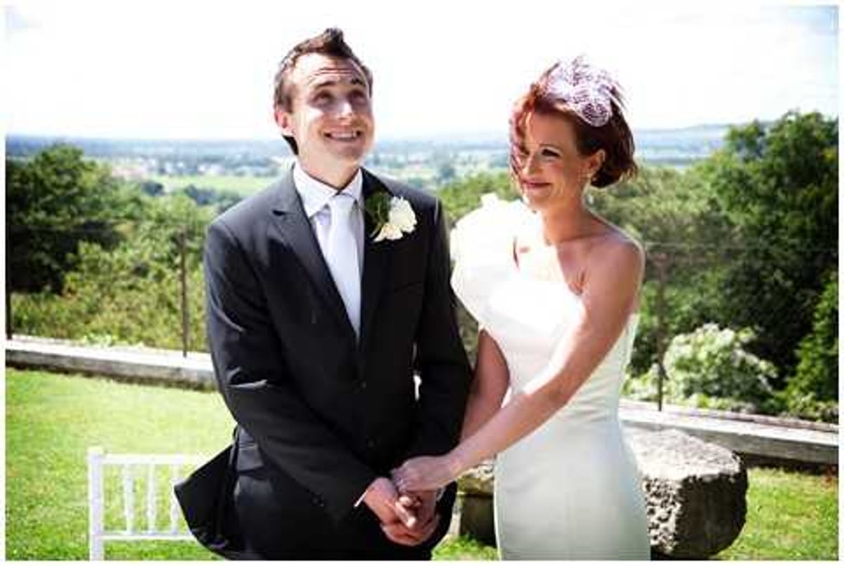A couple portrait taken outdoors on a terrace or garden area with a wide panoramic countryside view visible in the background. The groom wears a dark suit with a light blue tie and a white buttonhole flower, while the bride wears a fitted ivory strapless dress and a small pink fascinator in her red upswept hair. The couple are holding hands and laughing together, captured in a candid mid-shot. The styling is classic with a retro-inspired edge, reflected in the bride's hair accessory and sleek silhouette dress.