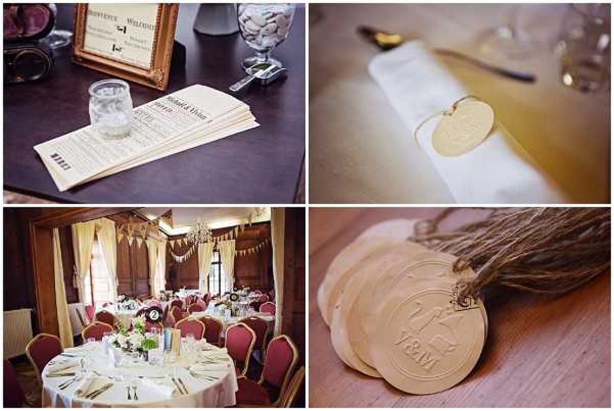 A four-image collage showing wedding reception details with a rustic, vintage styling theme. Top left: a close-up flat lay of printed wedding programs or menus in a newspaper-style format alongside a gold ornate frame, a glass jar filled with white candies, and a small glass vessel, arranged on a dark surface. Top right: a close-up detail shot of a white folded napkin tied with a natural twine bow and a small circular wooden tag, placed beside gold cutlery. Bottom left: a wide shot of an indoor reception room set for dinner, featuring round tables with white linens, red upholstered chairs, low floral centerpieces with green and white blooms, and bunting garlands in cream and warm tones hanging from a chandelier, with wood-paneled walls and draped curtain windows. Bottom right: a close-up flat lay of multiple circular wooden tags stamped with a monogram logo and couple's initials, tied together with natural jute twine, used as place card or favor tags.