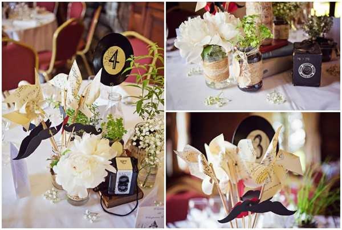 Three close-up detail shots of wedding reception table centerpieces arranged in a collage. The centerpieces feature a vintage-themed decor palette combining cream white peonies and baby's breath in mason jars wrapped with burlap ribbon, pinwheels made from sheet music paper, black cardboard mustache props on sticks, and vinyl record table numbers (numbers 3 and 4 visible) with gold lettering on black backgrounds. Small vintage-style toy cameras and stacked aged books are incorporated as decorative props. The overall styling theme is retro/vintage with a black, gold, cream, and kraft paper color palette. Tables are dressed with white linens and the reception appears to be held in an indoor ballroom or function room with red upholstered chairs visible in the background.