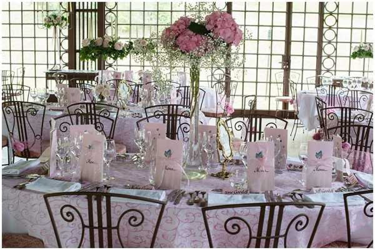 A reception tablescape detail shot taken indoors in what appears to be a glass-walled or windowed venue with decorative iron lattice panels. The round tables are dressed in pink patterned tablecloths and set with crystal glassware, silverware, and pink folded menu cards decorated with a small blue butterfly motif. Place cards are visible along the table edge. The centerpiece is a tall clear vase filled with pink hydrangeas and baby's breath. The chairs are dark wrought-iron with scrolled backs. The overall decor palette is soft pink and white with romantic, classic styling throughout the room.