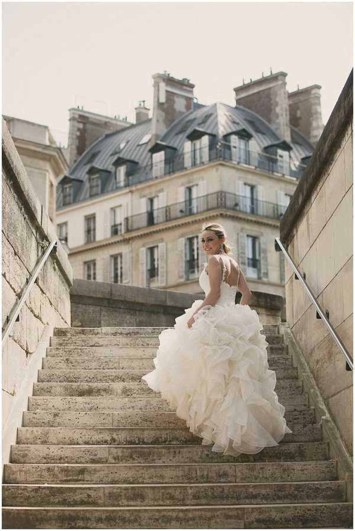 A bridal portrait of a single bride ascending a wide stone staircase with metal handrails, glancing back over her shoulder toward the camera. She wears an ivory ballgown with a heavily ruffled and layered organza skirt, a fitted lace bodice, and an open back with delicate strap detailing. The setting is outdoors in Paris, with a classic Haussmann-style building featuring a slate mansard roof, wrought-iron balconies, and tall shuttered windows visible in the background. The composition is a full-length portrait shot from a low angle, emphasizing the volume of the dress and the Parisian architectural backdrop.