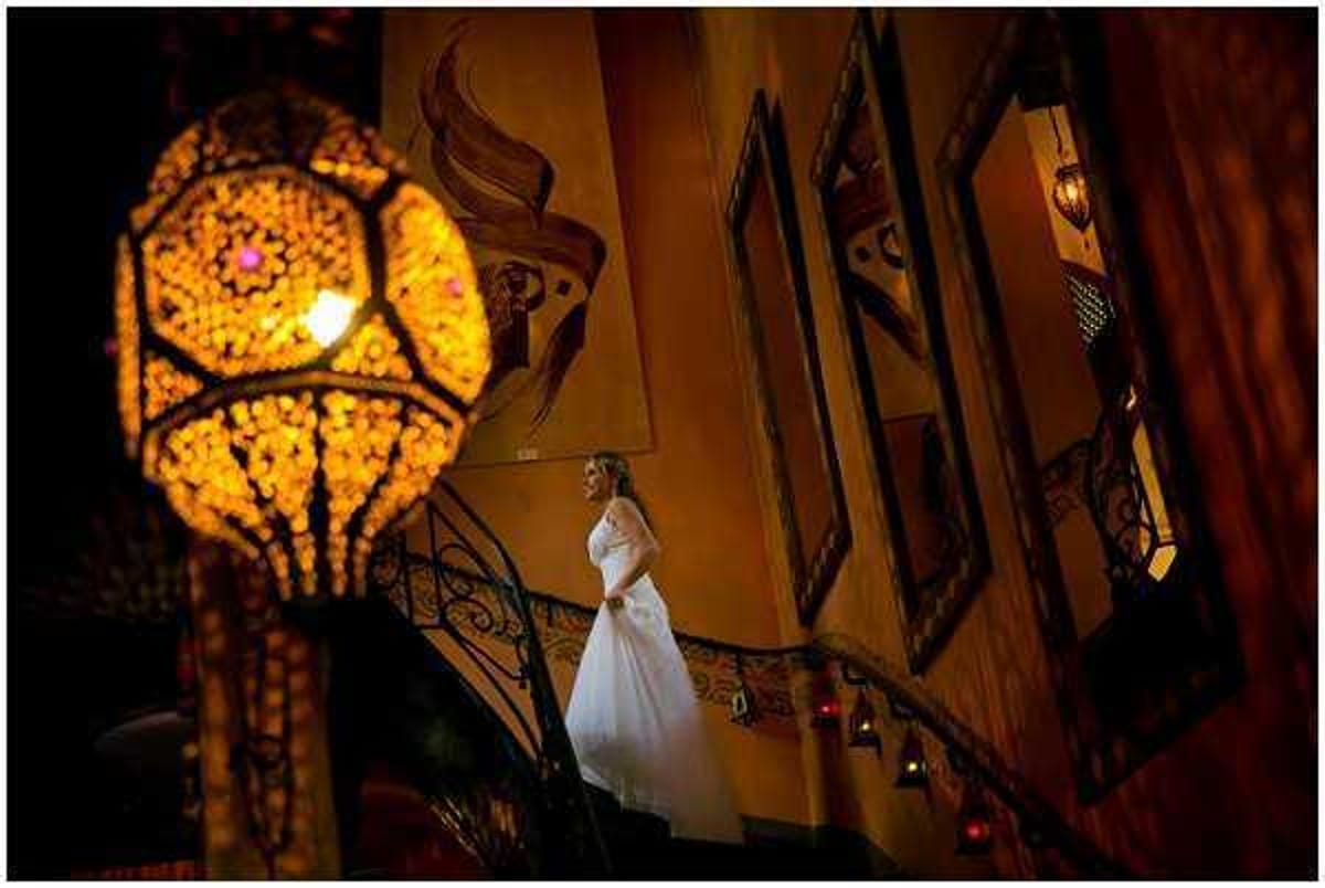 A bride in a long white gown with long sleeves stands on an ornate wrought-iron staircase inside a richly decorated interior, gazing to one side in a full-length portrait shot. The setting features warm amber-toned walls with decorative painted motifs, framed mirrors, and small red lanterns lining the staircase railing, suggesting a Moorish or riad-style venue. In the foreground, a large geometric mosaic lantern glows in deep amber and gold, dominating the left side of the frame and casting warm light throughout the scene. The overall decor palette is deep amber, gold, and red with an ornate, Middle Eastern-influenced aesthetic.
