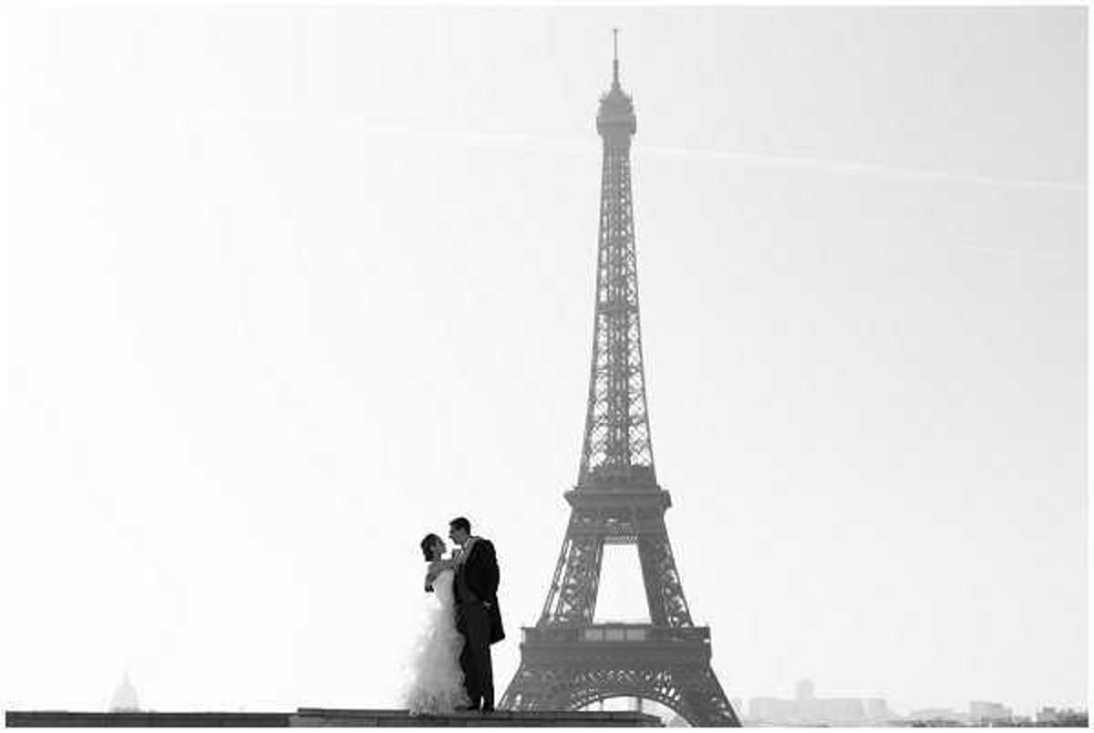 This black-and-white couple portrait shows a bride and groom sharing a kiss or close embrace outdoors in Paris, with the Eiffel Tower prominently centered in the background. The image is shot in high-key tones with a pale, overexposed sky creating strong contrast against the dark silhouette of the tower and the couple. The bride wears a full, ruffled ball gown with a layered skirt, and the groom is dressed in a dark suit. The wide shot is composed to place the couple small against the scale of the tower, emphasizing the iconic Parisian landmark as the dominant visual element.