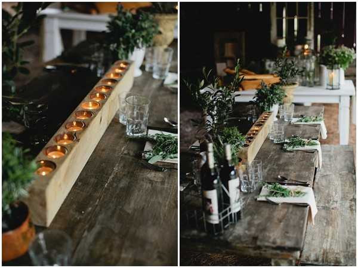 A diptych of reception table detail shots in a rustic indoor barn or farmhouse setting. The left image is a close-up of a raw wooden beam candle holder running the length of a reclaimed wood trestle table, with a row of lit tealight candles set into drilled holes and loose greenery scattered alongside glassware. The right image shows a wider view of the same table, revealing place settings with linen napkins, dark cutlery, glass tumblers, wine bottles in a wire caddy, and abundant fresh greenery including olive branches and herbs used as both centerpieces and plate garnish. The overall decor palette is dark and natural — raw wood, matte black accents, and deep green botanicals — consistent with a rustic or foraged styling theme with no flowers, relying entirely on candle warmth and greenery for decoration.