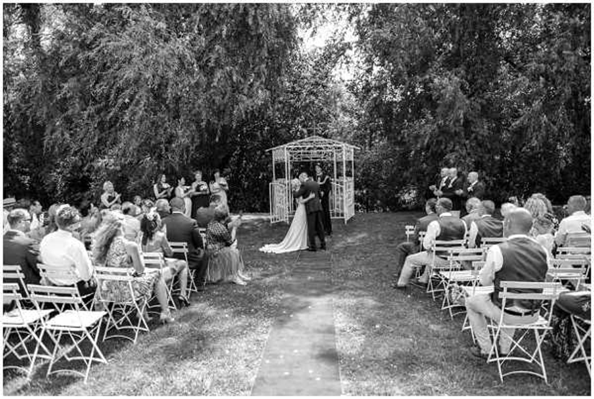 A black-and-white wide shot of an outdoor garden wedding ceremony, capturing the moment the couple shares their first kiss at the altar. The ceremony takes place on a grass lawn with a central aisle runner leading to a white metal gazebo-style arch where the couple and officiant are standing. Approximately 40–50 guests are seated in white folding chairs arranged in two sections on either side of the aisle, with a small bridal party visible to the left of the arch. The contrast is bright and airy, with dark foliage from large trees forming a natural backdrop behind the ceremony space.