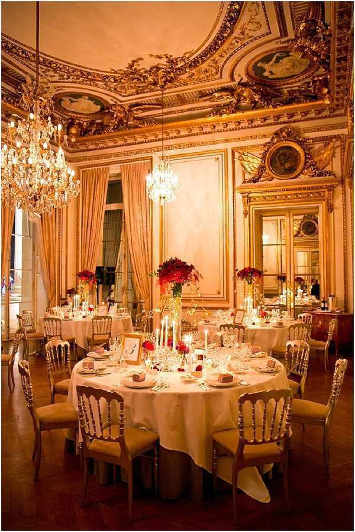 Indoor wedding reception setup in a grand Haussmann-era or palace-style ballroom featuring heavily gilded ceiling moldings, ornate painted medallions, large gold-framed mirrors, and crystal chandeliers casting warm amber light throughout the room. Round tables are dressed in ivory floor-length linens and set with gold Napoléon III-style chairs with tan upholstered seats. Centerpieces consist of tall and low arrangements of deep red flowers, likely red dahlias or red roses, paired with white taper candles in varying heights. Place settings include crystal glassware, white napkins, and small table number frames. The decor palette is rich red, ivory, and gold, giving the room a classic formal style. Wide-angle shot taken from a low perspective, showing multiple tables arranged across the reception space. Potential venue feature image.