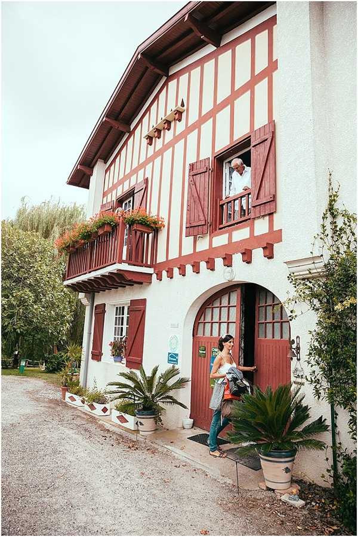 A wide exterior shot of a traditional Basque-style two-storey building featuring white render with deep red timber framing, red wooden shutters, and a balcony with flower boxes planted with orange and red blooms. A woman in casual clothing — an orange top and jeans — stands at the arched front door, while an older man leans out of an upper-floor window. Potted palm plants and climbing greenery flank the entrance. The setting is a gravel courtyard in front of the building. This appears to be a pre-wedding arrival or venue visit moment rather than a formal wedding event. Potential venue feature image.