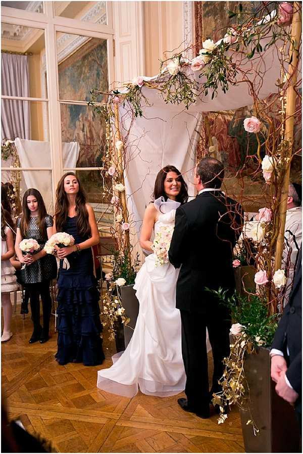 An indoor wedding ceremony taking place in a grand room with ornate painted murals on the walls and a parquet wood floor, consistent with a French chateau or historic mansion. The bride, wearing a white sleeveless high-neck gown, faces the groom who is dressed in a black tuxedo, and she holds a cascading bouquet of ivory and blush flowers. They stand beneath a tall ceremony arch constructed from twisted branches and tall wooden posts, decorated with blush and ivory roses, trailing greenery, and hanging curly willow branches. Two bridesmaids stand to the left — one in a navy tiered ruffle gown and one in a navy sequined dress — with a small guest in a silver sequined top also visible; a bouquet of ivory and cream roses is held by one of the bridesmaids. The ceremony decor features a romantic, organic style with the warm-toned branch structure, soft pink and white floral accents, and a white fabric drape as a backdrop. The shot is a medium wide portrait-style image capturing the couple and the immediate bridal party during the exchange of vows.