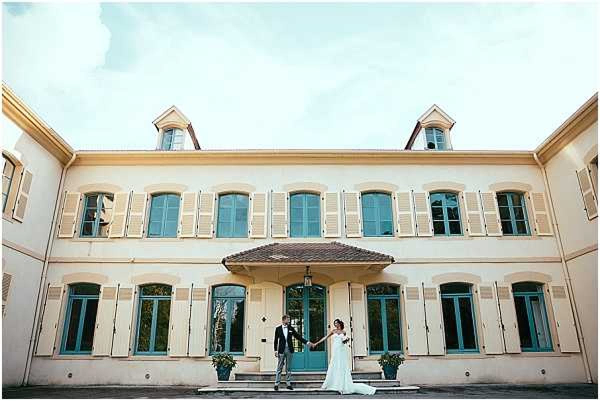 A couple poses hand-in-hand in front of a large two-story French manor house or bastide, standing at the base of the main entrance steps. The bride wears a white strapless gown with a train, and the groom wears a dark navy suit. The building features cream-rendered facades with teal-painted shutters and arched windows, a central covered entrance with teal double doors, and dormer windows on the upper floor. The shot is a wide portrait composition that places the couple small against the full width of the building, emphasizing the architecture. Potential venue feature image.