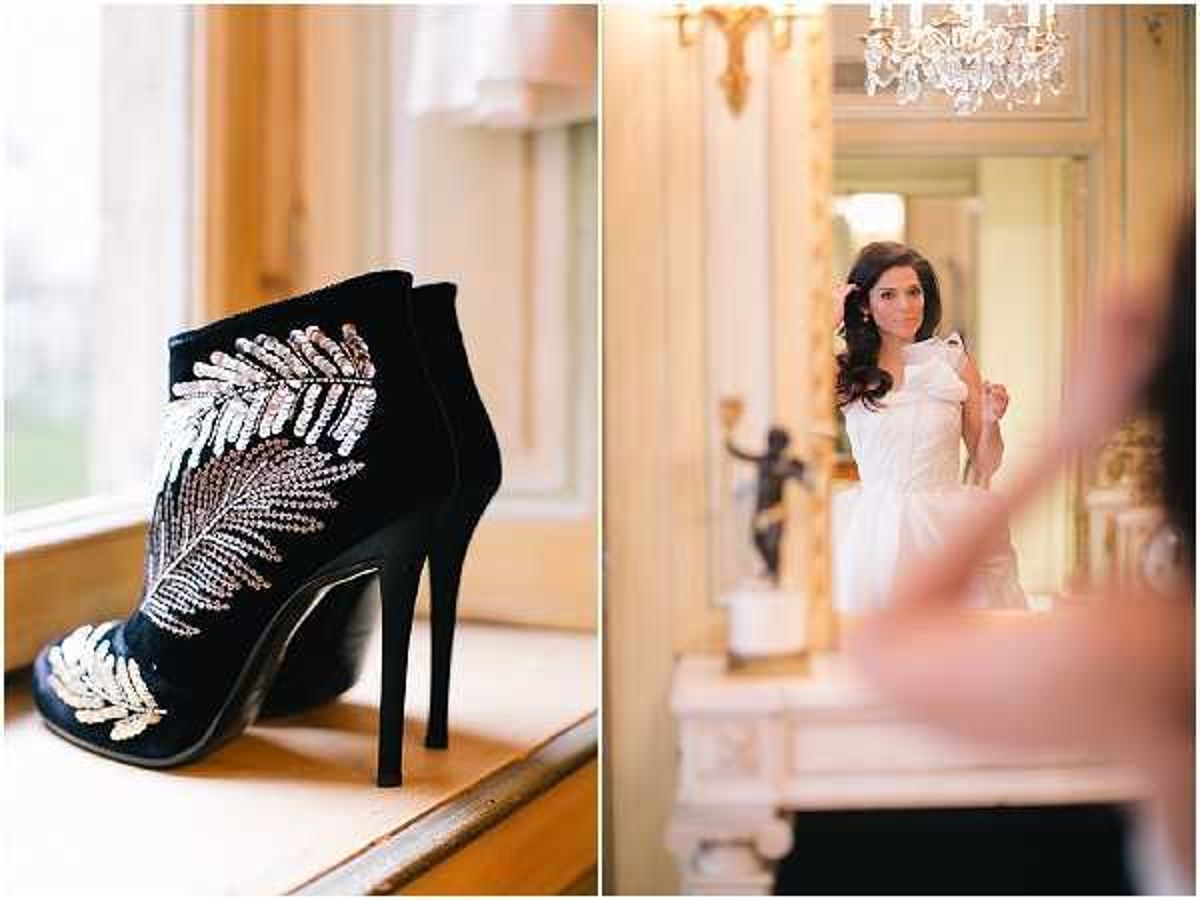 A diptych of two getting-ready images. On the left, a close-up detail shot of a black stiletto ankle boot decorated with silver and gold crystal beading in a feather motif, placed on a wooden windowsill with natural light behind it. On the right, a bride in a white strapless structured ball gown with a large bow detail stands in an ornate interior room, viewed through a mirror reflection; the room features a crystal chandelier, gilded mirror frames, a white carved mantelpiece, and a dark bronze figurine statue, suggesting a classic French chateau or period venue. The bride has long dark wavy hair and is being assisted by another person partially visible in the foreground. The overall styling is classic and formal.
