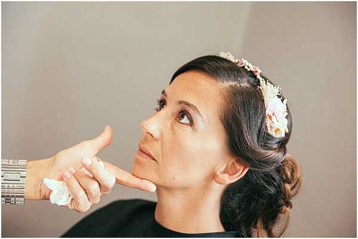 A close-up portrait of a bride during her getting-ready preparations, with a makeup artist's hand visible applying or blending product to her chin area. The bride is looking upward with her eyes directed toward the ceiling, wearing a black makeup cape. Her dark hair is styled in a low updo adorned with a floral hair accessory featuring small peach and blush flowers with dried or delicate blooms. The makeup artist wears a wide silver cuff bracelet. The background is a plain neutral grey, suggesting an indoor preparation room.