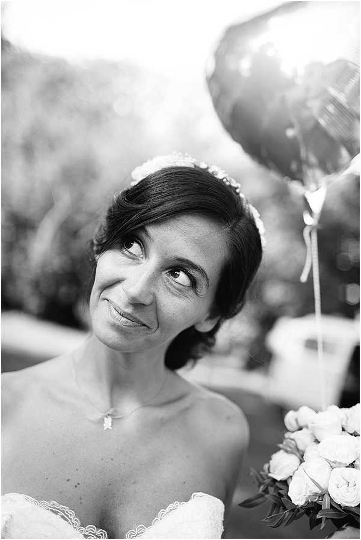 A black-and-white close-up portrait of a bride outdoors, glancing upward with a slight smile. She wears an off-the-shoulder lace gown and a floral headpiece, with a delicate pendant necklace. In her hand she holds a bouquet of what appear to be garden roses with foliage, and a heart-shaped foil balloon is visible in the background to her right. The background is heavily blurred with soft bokeh, keeping the focus on her face. The image has strong contrast with deep mid-tones and bright highlights.