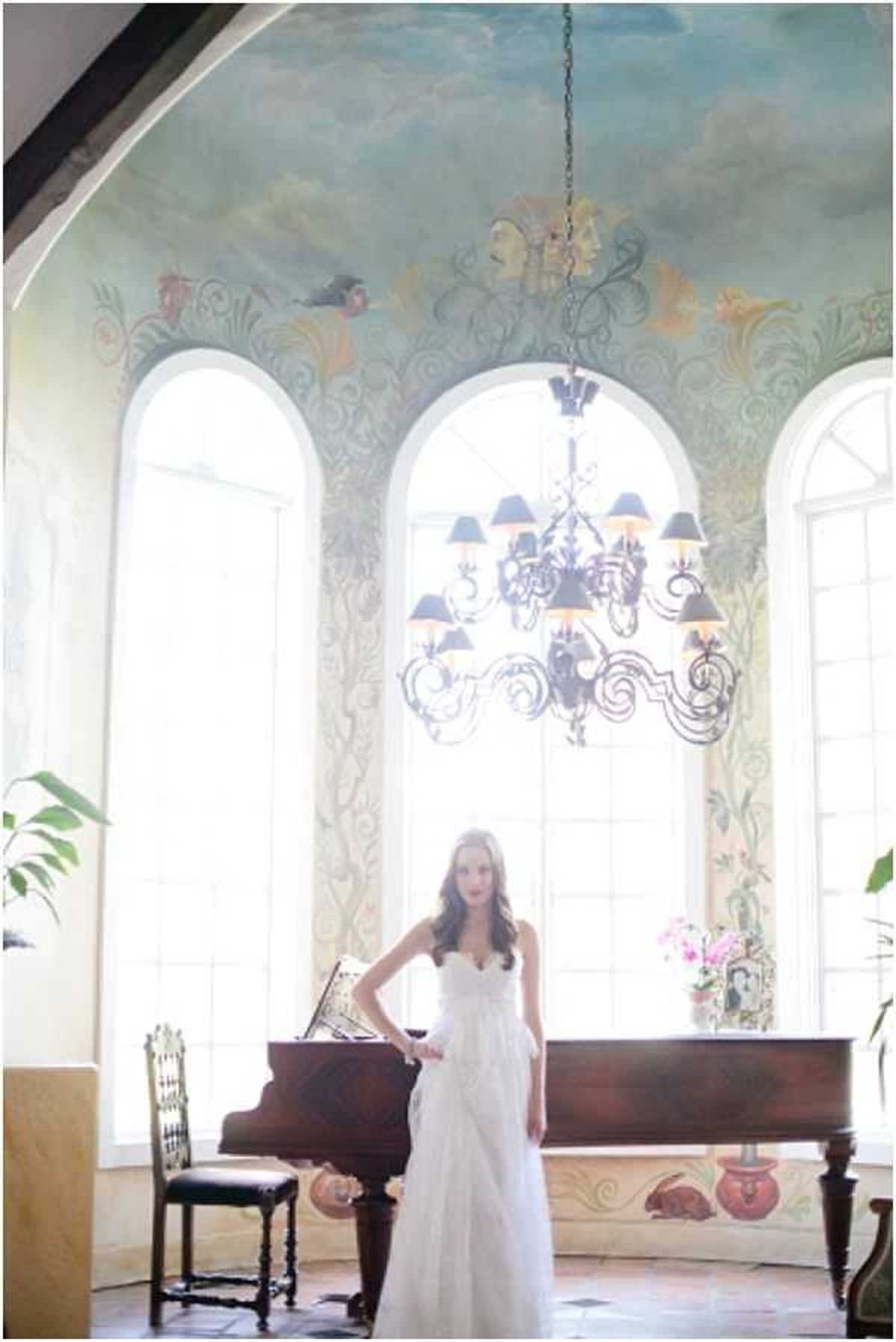 A bridal portrait of a single bride standing indoors in front of a grand piano in a room with floor-to-ceiling hand-painted murals covering both the walls and ceiling, depicting clouds, figures, birds, and ornate scrollwork in soft blues, greens, and gold tones. The bride wears a strapless white lace A-line gown with a sweetheart neckline and poses with one hand on her hip. A wrought-iron chandelier with black lampshades hangs centrally from the painted ceiling, and three tall arched windows backlight the scene. The room also contains a carved wooden chair and a pink floral arrangement on the piano. The composition is a full-length portrait shot with the architectural mural as a prominent backdrop. Potential venue feature image.