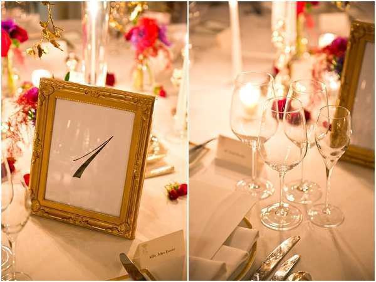 Close-up detail shots of a wedding reception table setting, shown as a diptych. The left image features a gold ornate picture frame displaying the table number '1' in black calligraphy script, surrounded by vibrant fuchsia, red, and magenta floral arrangements, white taper candles in gold holders, and place cards on a white linen tablecloth. The right image shows the same table from a different angle, highlighting multiple crystal wine glasses and a champagne flute, with a lit votive candle, folded white napkins with silverware, and a gold-framed element visible in the background. The decor palette combines gold accents with bold jewel-toned florals, suggesting a formal indoor reception with a classic yet vivid color scheme.