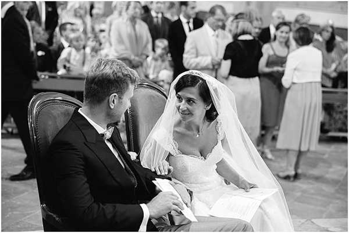 A black-and-white image of a bride and groom seated side by side during an indoor wedding ceremony, likely in a chapel or formal hall. The bride, wearing a strapless lace-bodiced gown and a long veil with lace trim, turns to look at the groom with a warm smile while holding papers, possibly vows or an order of service. The groom wears a dark suit with a boutonniere. In the soft background, approximately 15–20 guests are visible standing and conversing, suggesting the ceremony is either just beginning or concluding. The image is a medium close-up portrait with high contrast tones, keeping the couple sharp against a naturally blurred crowd.