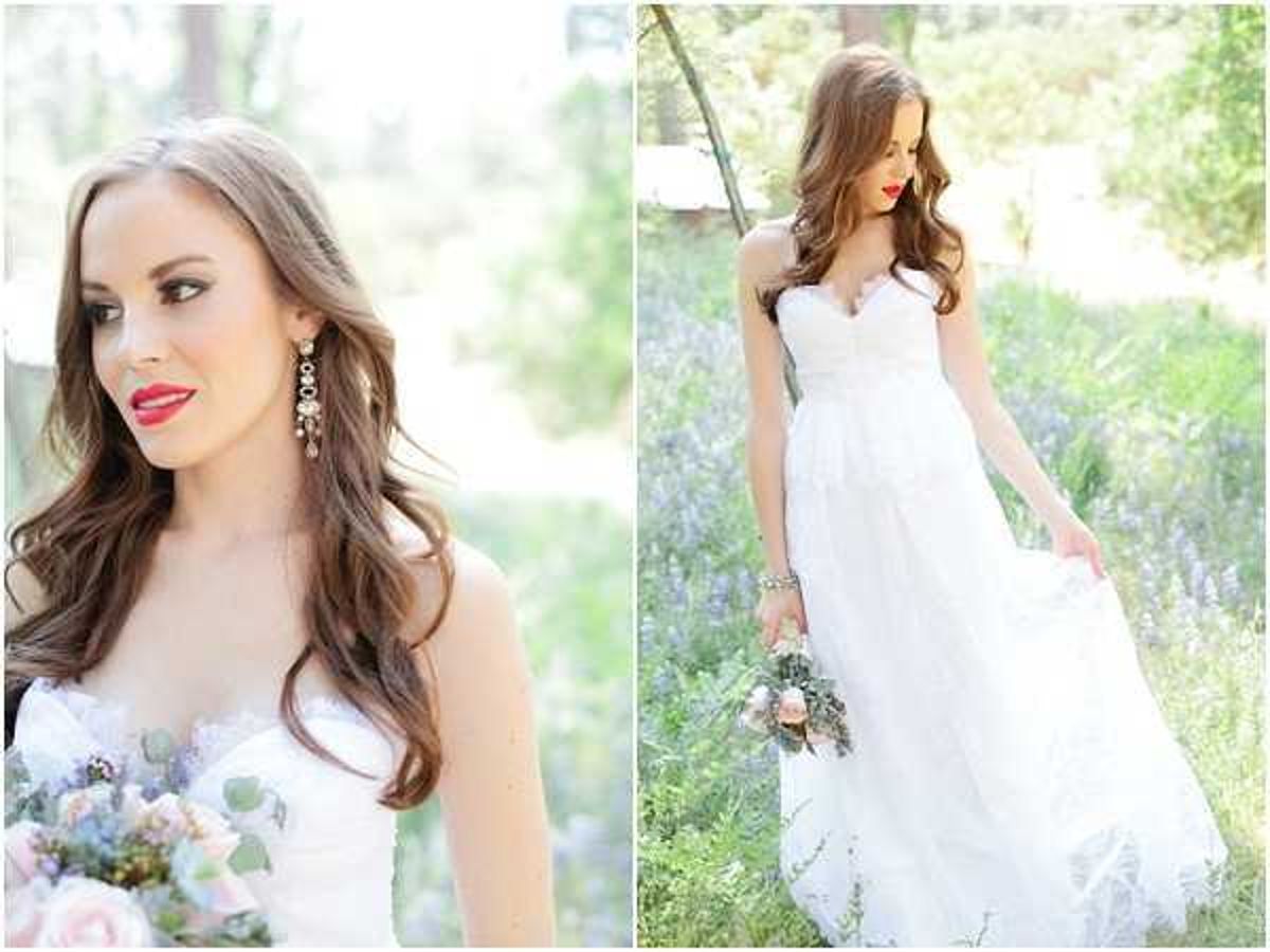A bridal portrait diptych showing the same bride in two compositions: a close-up on the left and a full-length shot on the right. The bride wears a strapless sweetheart-neckline white gown with a full skirt and lace detailing, and holds a bouquet of blush pink roses and eucalyptus. She has long wavy brown hair worn down, bold red lipstick, and drop crystal earrings. In the full-length shot, she stands outdoors among tall lavender-purple wildflowers and lifts the skirt of her dress slightly. The styling combines a classic white gown with a bold makeup look. Both shots are bright and softly lit with a light, airy quality.