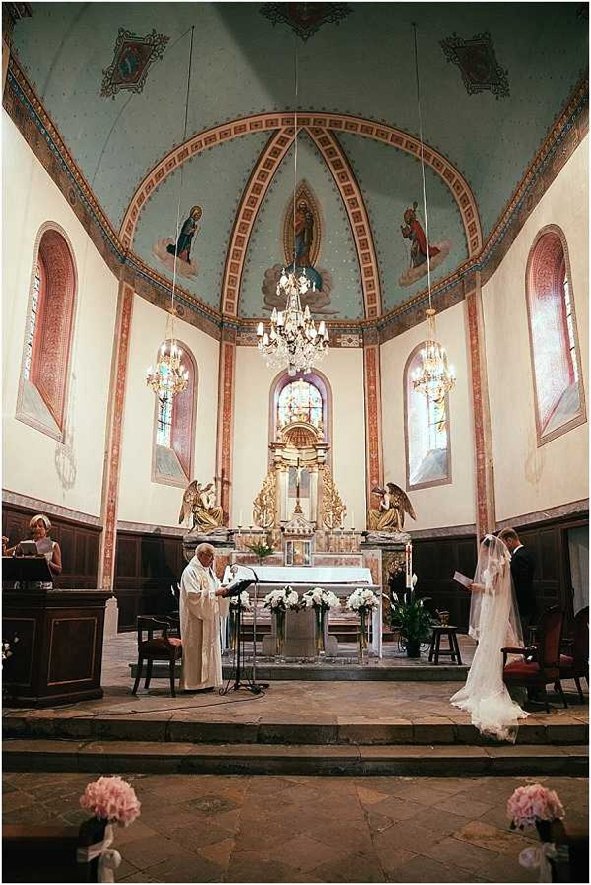A Catholic wedding ceremony taking place inside a French chapel with a painted vaulted apse ceiling in pale blue and terracotta tones, decorated with religious frescoes and gilt detailing. A priest in white robes reads at a lectern before the ornate gold altar, while the bride in a white gown with a long cathedral veil stands to the right facing him; the groom is partially visible beside her. The altar is flanked by arrangements of white flowers, likely roses and lilies, in tall vases, and crystal chandeliers hang from the ceiling. Pink peony bouquets tied with white ribbon are attached to the ends of the stone pews in the foreground, and stained glass windows frame the apse on either side. Wide shot taken from the nave looking toward the altar, capturing the full architectural interior of the chapel. Potential venue feature image.