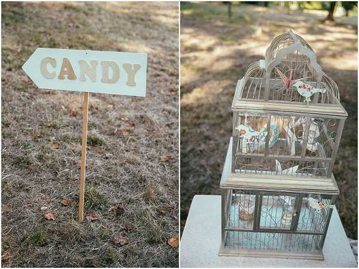 A close-up detail shot composed of two side-by-side images showing rustic wedding decor elements in an outdoor setting. On the left, a white wooden arrow-shaped sign reading 'CANDY' in cut-out lettering is staked into the ground on a wooden dowel. On the right, a vintage-style white metal birdcage displayed on a white pedestal contains decorative paper elements, small floral accents in coral and white tones, and what appears to be butterfly decorations. The overall styling is rustic-vintage with a soft, muted color palette. No people are visible in either frame.