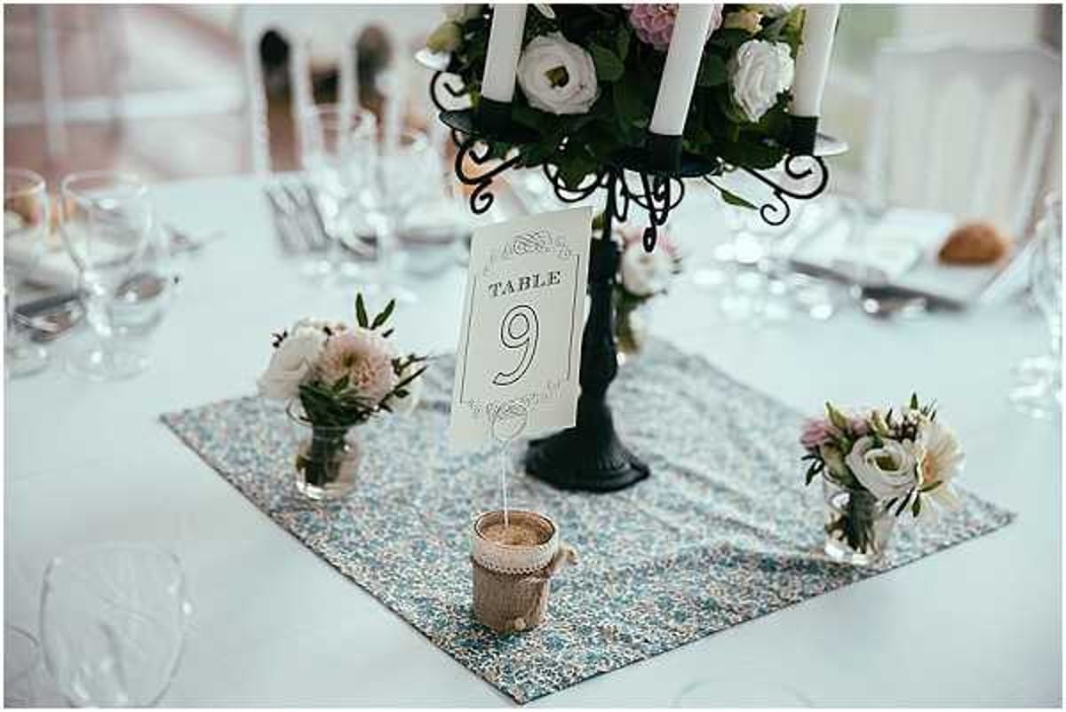 Close-up detail shot of a wedding reception table centerpiece setup. The table features a black wrought-iron candelabra holding white taper candles and topped with white lisianthus and blush pink flowers with greenery. A ornate-bordered table number card reading 'Table 9' is displayed in a small burlap-wrapped holder. Two small bud vases contain clusters of blush pink and cream flowers, including what appear to be lisianthus and small spray roses. These elements are arranged on a Liberty-print floral fabric table runner in muted blue and pink tones, laid over a white tablecloth. Crystal glassware and white place settings are visible in the soft-focus background. The overall decor palette is blush pink, cream, and black with a vintage-romantic styling theme.