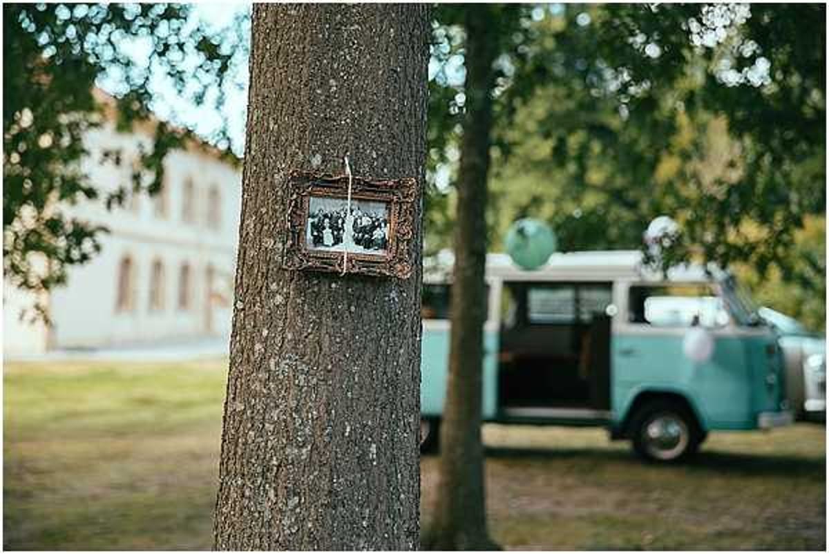 An outdoor wedding decor detail shot showing a small rustic wooden frame with a black-and-white photograph hanging on a tree trunk, likely a vintage family photo used as sentimental decor. In the background, softly out of focus, is a mint green vintage Volkswagen camper van with its side door open, decorated with a large mint balloon, serving as a wedding prop or photo booth station. A light-colored venue building is visible in the far background. The overall styling theme is rustic-vintage with a mint green color accent.