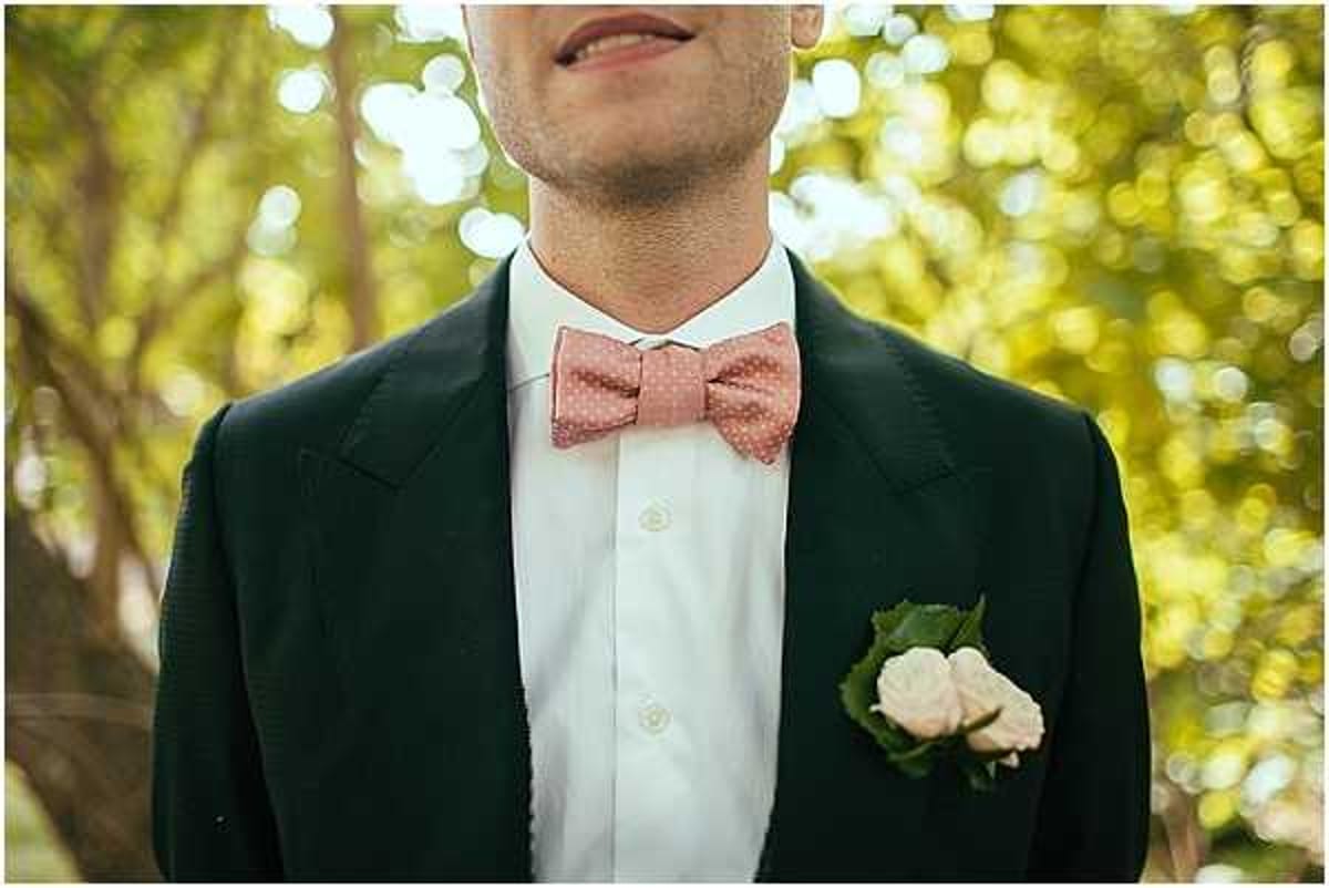 Close-up portrait of a groom from chin to waist, wearing a dark navy suit jacket with a white dress shirt and a dusty rose polka-dot bow tie. His pocket square is replaced by a boutonniere featuring a small cream rose bud with green foliage tucked into the breast pocket. The background is soft and blurred with green foliage, suggesting an outdoor garden or wooded setting. The styling has a playful yet classic feel, with the patterned bow tie as a deliberate style accent.