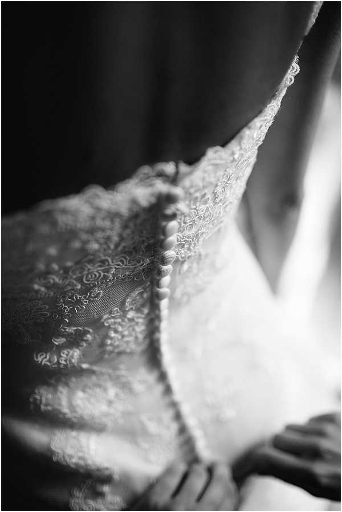 A black-and-white close-up detail shot of a bride being helped into her wedding dress during the getting-ready portion of the day. The image focuses on the back of the gown, which features intricate floral lace fabric and a column of small fabric-covered buttons running down the center back. A second person's hands are partially visible at the lower right, assisting with fastening the buttons. The composition uses shallow depth of field with soft window light from the right, creating strong contrast between the bright white lace and the dark background.