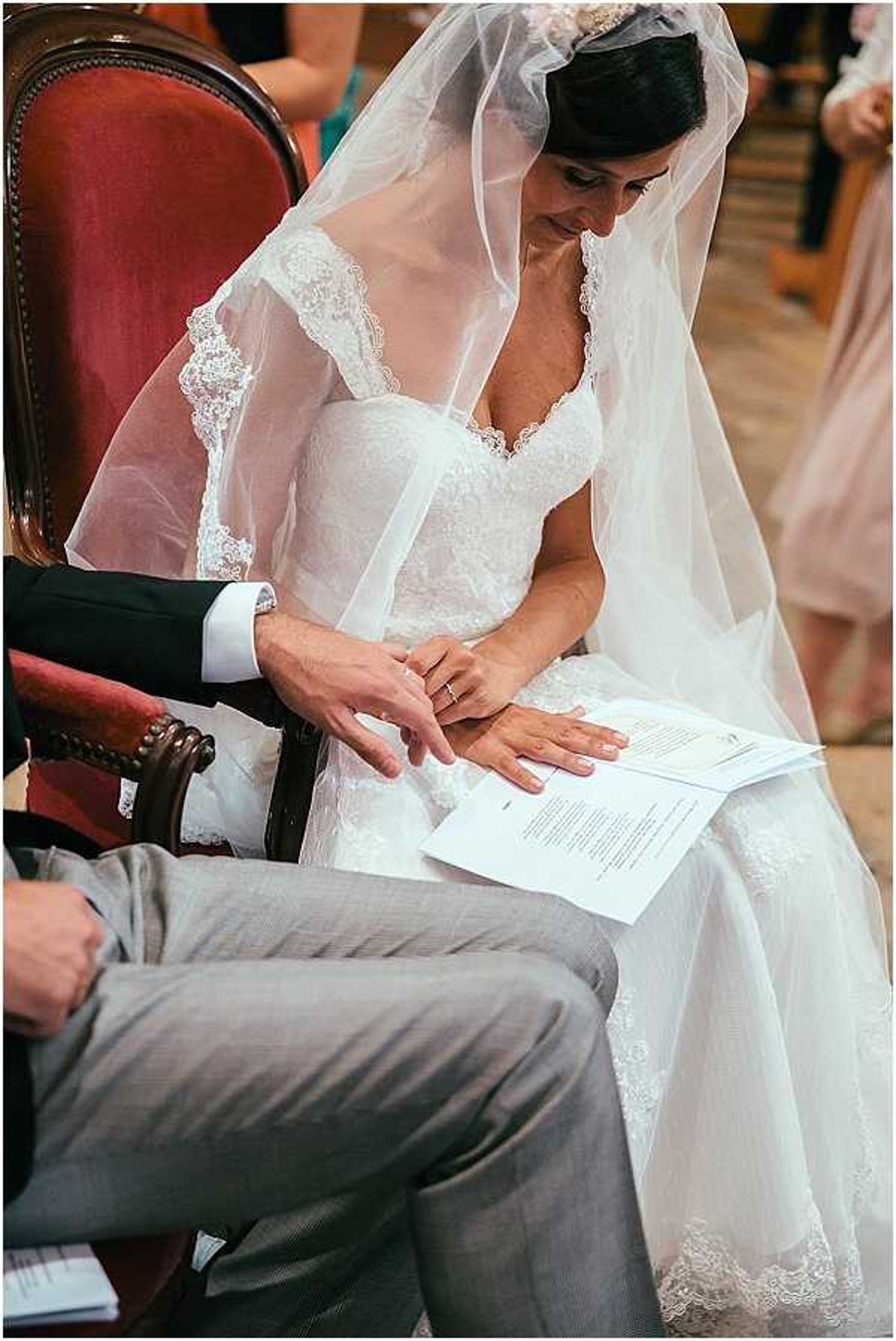 A close-up portrait shot of a bride seated during an indoor religious ceremony, looking down at a printed ceremony booklet resting on her lap. She wears a white lace gown with a sweetheart neckline and lace-trimmed long veil, along with a small tiara headpiece. The groom, seated beside her in grey trousers and a dark jacket, reaches over to touch her hand on the booklet. The ornate wooden chairs with red upholstery suggest a church or chapel setting. Bridesmaids in blush pink dresses are partially visible in the background.