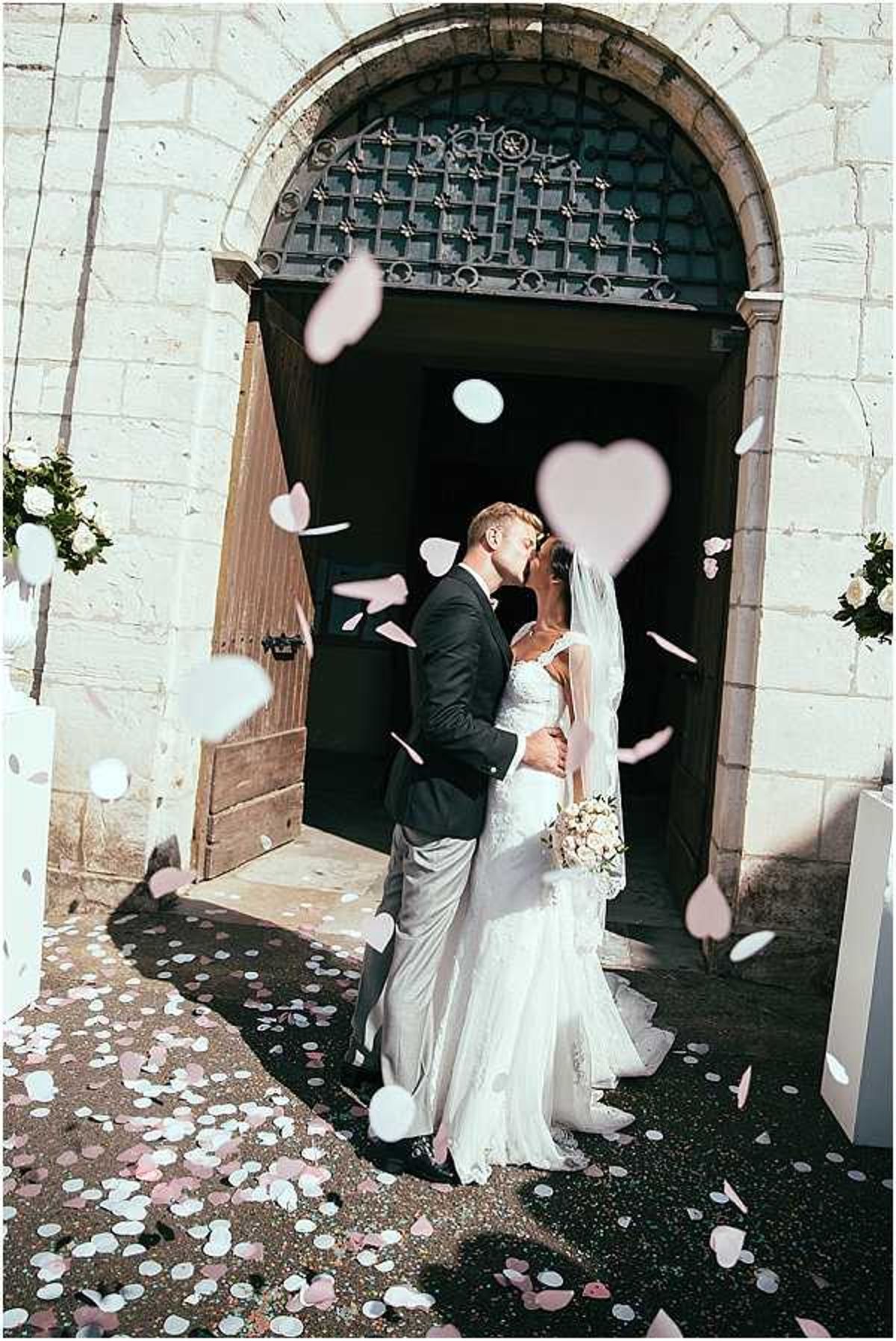 The bride and groom share a kiss on the steps outside a stone chapel immediately following their ceremony, with pink and white heart-shaped and round confetti pieces filling the air and scattered across the ground around them. The bride wears a fitted, lace wedding gown with a long veil and carries a bouquet of blush and ivory blooms, while the groom wears a dark navy blazer with grey trousers. Floral arrangements of white flowers and greenery are visible on either side of the arched chapel doorway, which features ornate dark ironwork. The shot is a full-length portrait framing the couple centrally against the chapel entrance, with confetti pieces in the foreground creating a layered, celebratory effect.