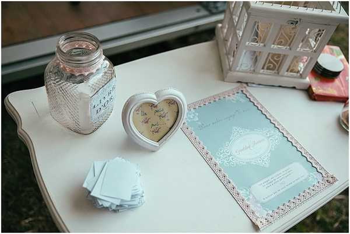 Close-up detail shot of a wedding guest book station arranged on a small white vintage side table. The display includes a glass mason jar labeled 'Love' filled with folded notes or coins, a small white heart-shaped photo frame containing a handwritten message, a stack of pale blue guest cards for writing wishes, and a pale blue wedding program or guest book with pink lace-style border detailing. A white ornamental birdcage lantern sits in the background along with what appear to be small decorative items. The overall decor palette is pale blue, white, and soft pink, consistent with a romantic vintage or shabby-chic styling theme.