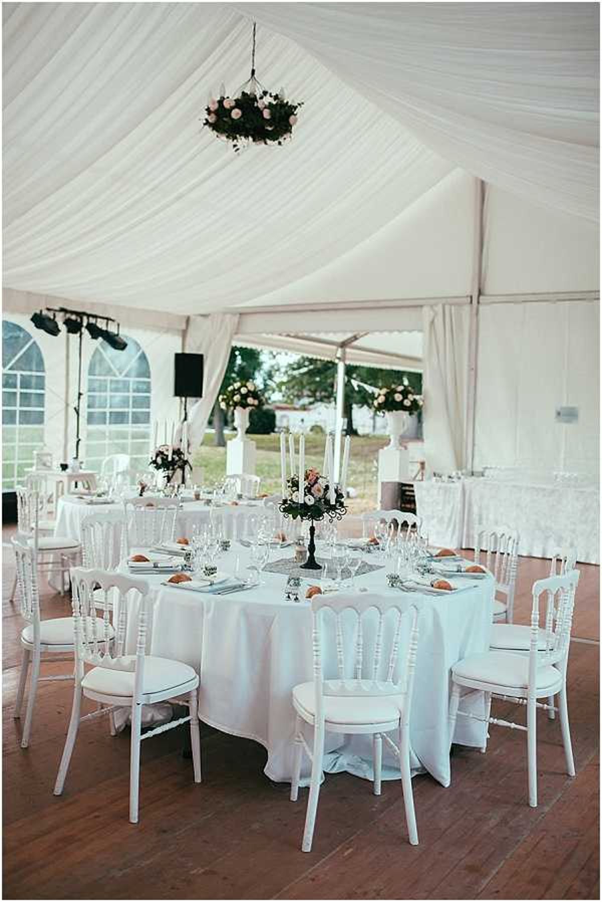 A wide shot of a wedding reception setup inside a white draped marquee tent with a warm wooden floor. The foreground features a round table dressed in a white floor-length linen, surrounded by white Napoleon chairs with white cushioned seats, set for approximately eight guests with glassware, cutlery, and small orange bread rolls at each place setting. The centerpiece is a black candelabra holding tall white taper candles and a small arrangement of blush pink roses and greenery. A hanging floral chandelier with deep green foliage and blush blooms is suspended from the white ceiling draping above. Additional round tables with matching white linens and white chairs are visible in the background, along with tall white urn floral arrangements of blush roses positioned near the marquee's open sides. The overall decor palette is white and blush pink with black metallic accents, giving the space a classic, formal feel.