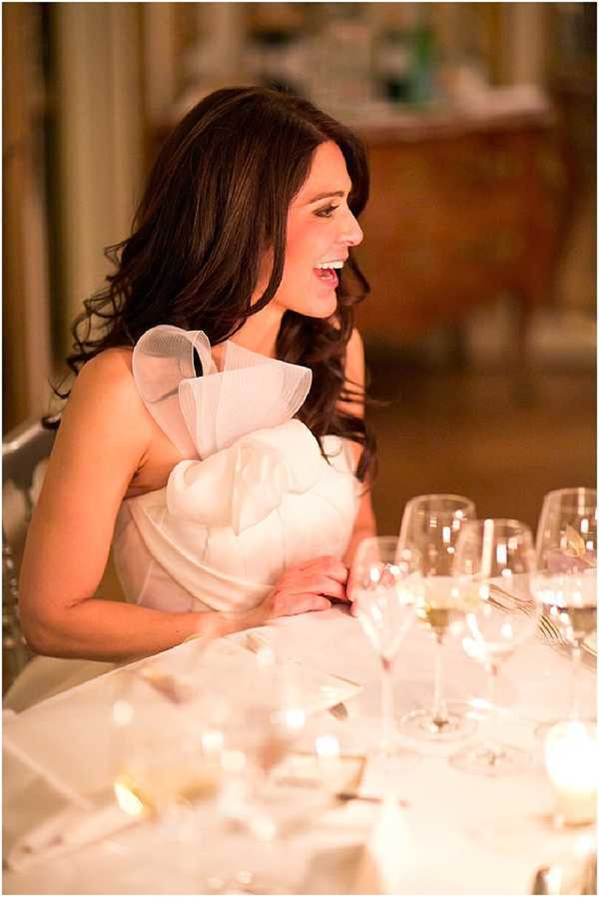 A bride is seated at what appears to be the head table during an indoor wedding reception, laughing and looking to one side. She is wearing a white one-shoulder gown with a large structured organza ruffle detail at the neckline and chest. Her dark brown hair falls in loose waves over her shoulders. The table in front of her is set with white linens, crystal wine glasses, and small votive candles providing warm ambient lighting. The background is softly blurred, revealing ornate gilded furniture consistent with a chateau or formal ballroom setting. This is a mid-range portrait shot with warm candlelit tones.