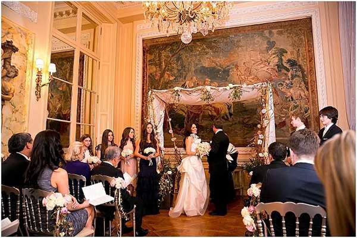 An indoor wedding ceremony taking place in a formal salon of what appears to be a French chateau or historic mansion, featuring ornate gold molding, a crystal chandelier, and a large decorative tapestry covering the back wall. The bride wears a full-skirted blush/ivory ballgown and holds a white floral bouquet, standing opposite the groom in a black tuxedo beneath a fabric chuppah-style canopy draped in ivory cloth and decorated with white and green floral arrangements on its four posts. The bridal party includes approximately three bridesmaids in dark navy floor-length gowns positioned to the left, while around 15-20 seated guests in formal attire watch the ceremony from black Chiavari chairs. The decor palette is classic and formal — black, ivory, and gold — with white and cream florals accenting the ceremony arch and chair arrangements, fitting a classic, formal styling theme. Wide-angle shot capturing the full ceremony scene. Potential venue feature image.