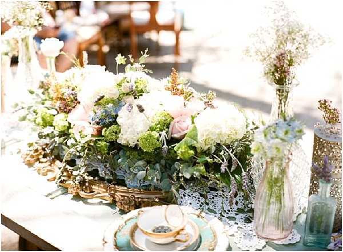 A close-up detail shot of an outdoor wedding reception tablescape styled in a vintage, romantic aesthetic. The centerpiece features a low, lush floral arrangement in an ornate gold-toned tray, comprising white hydrangeas, blush roses, green viburnum, eucalyptus, and small blue accent flowers. Surrounding the centerpiece are slender pink glass bud vases holding baby's breath and dried lavender-toned stems, along with a decorative blue-green glass bottle. The place settings include mismatched vintage china teacups and saucers in mint green and gold, layered on lace doilies over a white tablecloth. Wooden chairs and additional floral arrangements are visible in the softly blurred background, suggesting a sunny outdoor setting.