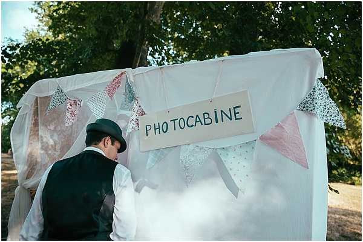A man wearing a dark waistcoat and a black bowler hat is bent over, appearing to set up or adjust a DIY photo booth at an outdoor wedding reception. The booth is constructed from a white fabric backdrop decorated with handmade fabric bunting in pastel tones including dusty pink, mint green, and floral patterned triangles. A hand-lettered cardboard sign reading 'PHOTOCABINE' hangs at the top center of the structure. The shot is a medium wide-angle taken from behind the man, set against a tree-lined outdoor venue. The overall styling is rustic and whimsical with a DIY aesthetic.