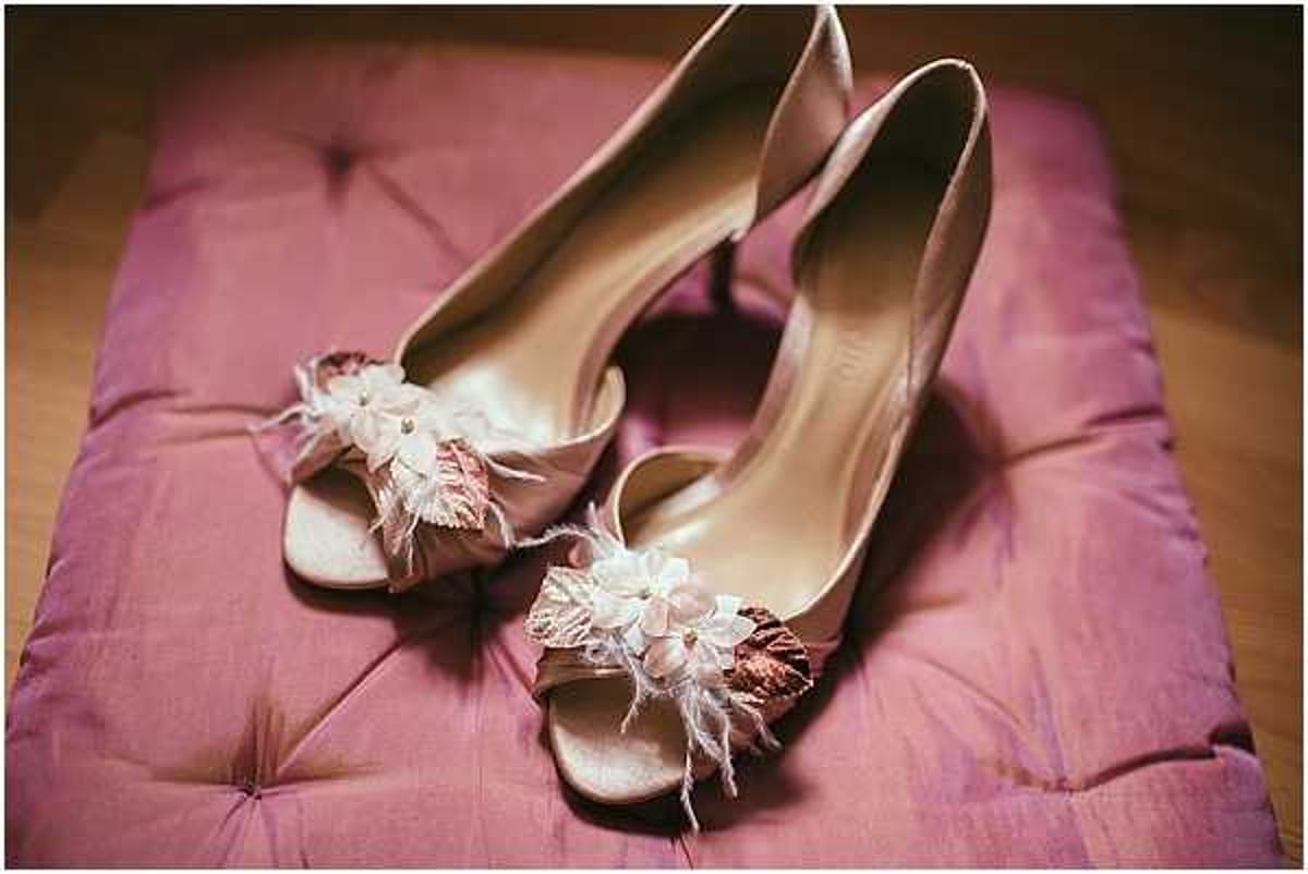 Close-up detail shot of a pair of bridal heels placed on a tufted mauve/dusty pink cushion or ottoman. The shoes are open-toe kitten heels in a champagne/blush satin fabric, each adorned with a decorative clip featuring small white fabric flowers, feather trim, and rose gold or copper-toned leaf embellishments. The styling is romantic and vintage-inspired. The warm ambient lighting gives the image a soft, slightly golden tone.