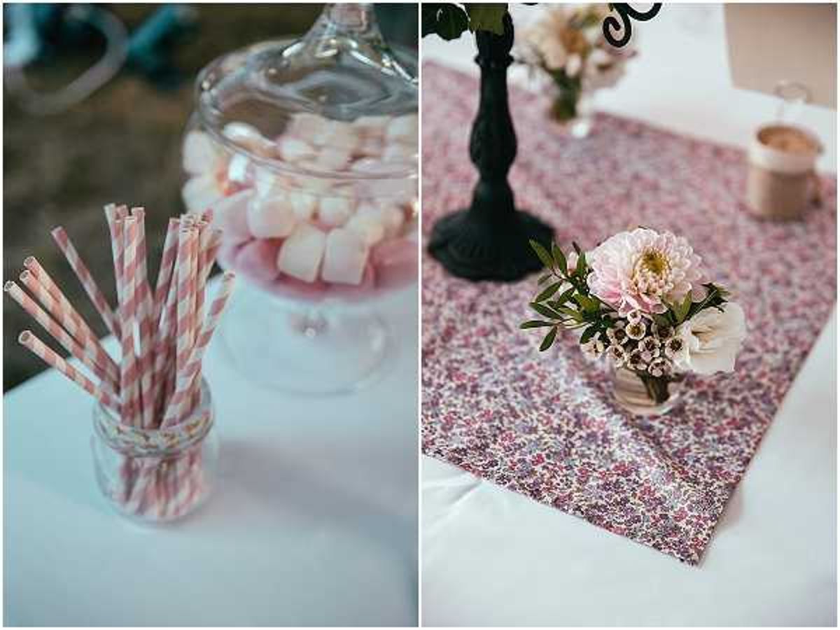 A split detail shot showing two reception table styling elements. On the left, a small glass jar holds pink and white striped paper straws alongside a glass apothecary jar filled with pink and white marshmallows on a white linen surface. On the right, a small glass vase holds a cluster of pink dahlias, white waxflower, and green foliage, placed on a burgundy and pink floral-print fabric table runner over a white tablecloth, alongside a black candelabra base and a small gold vessel in the background. The overall decor palette is soft pink, burgundy, and ivory with a vintage or garden-party styling theme. Close-up detail composition.