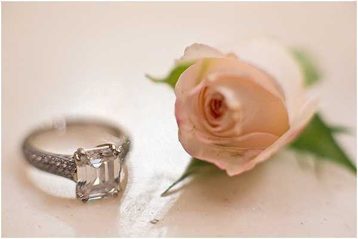 A close-up detail shot of a silver engagement ring featuring a large emerald-cut diamond solitaire with a pavé-set band, placed on a soft white surface beside a single blush pink rose boutonniere with green foliage. The shallow depth of field keeps the ring in sharp focus while the rose is slightly soft in the background. The overall palette is pale and muted, with warm ivory tones in the surface and soft pink in the rose.