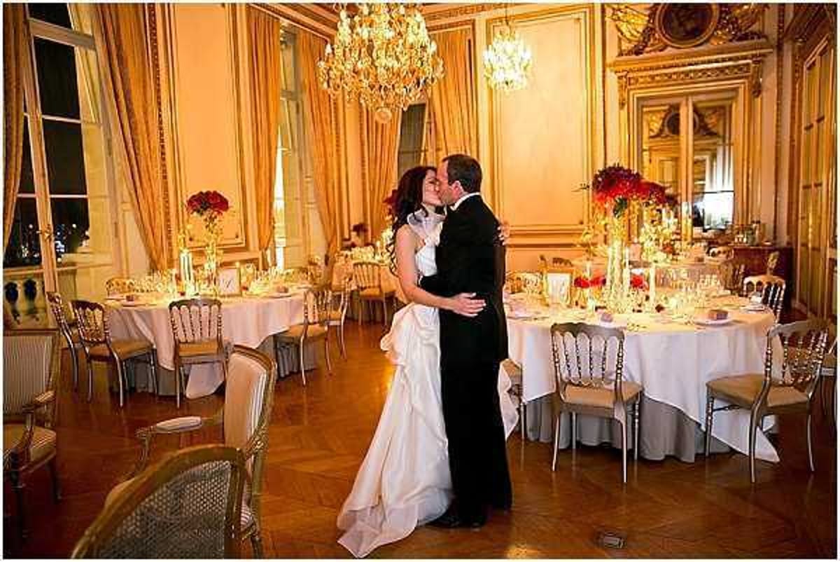 A bride and groom share a kiss on the dance floor of an ornate indoor ballroom, likely a Parisian hôtel particulier or grand reception venue. The room features gold-paneled walls, crystal chandeliers, and parquet flooring, with round dining tables set with white floor-length linens, candles, and bold red floral centerpieces. The bride wears a white strapless ball gown with a full skirt and a white fur or feather wrap around her shoulders, while the groom is dressed in a classic black tuxedo. The overall decor palette is gold, white, and deep red, creating a formal, classic aesthetic. Wide portrait shot capturing the couple centrally framed against the fully dressed reception room. Potential venue feature image.