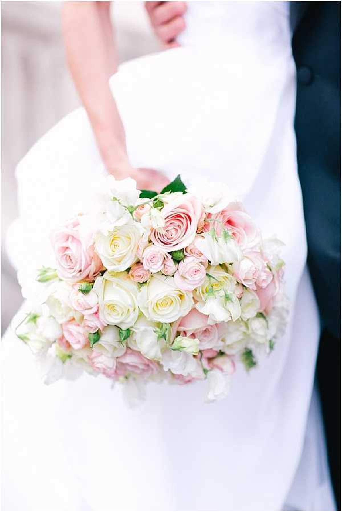 Close-up detail shot of a bridal bouquet held by the bride, with the groom partially visible in the background wearing a dark navy suit. The bouquet is a rounded, densely arranged composition of blush pink roses, cream roses, small spray roses in pale pink, and what appear to be white lisianthus blooms, with touches of green foliage. The bride is wearing a white gown with a smooth, fitted silhouette. The overall color palette is soft blush and cream, consistent with a classic, romantic styling approach.