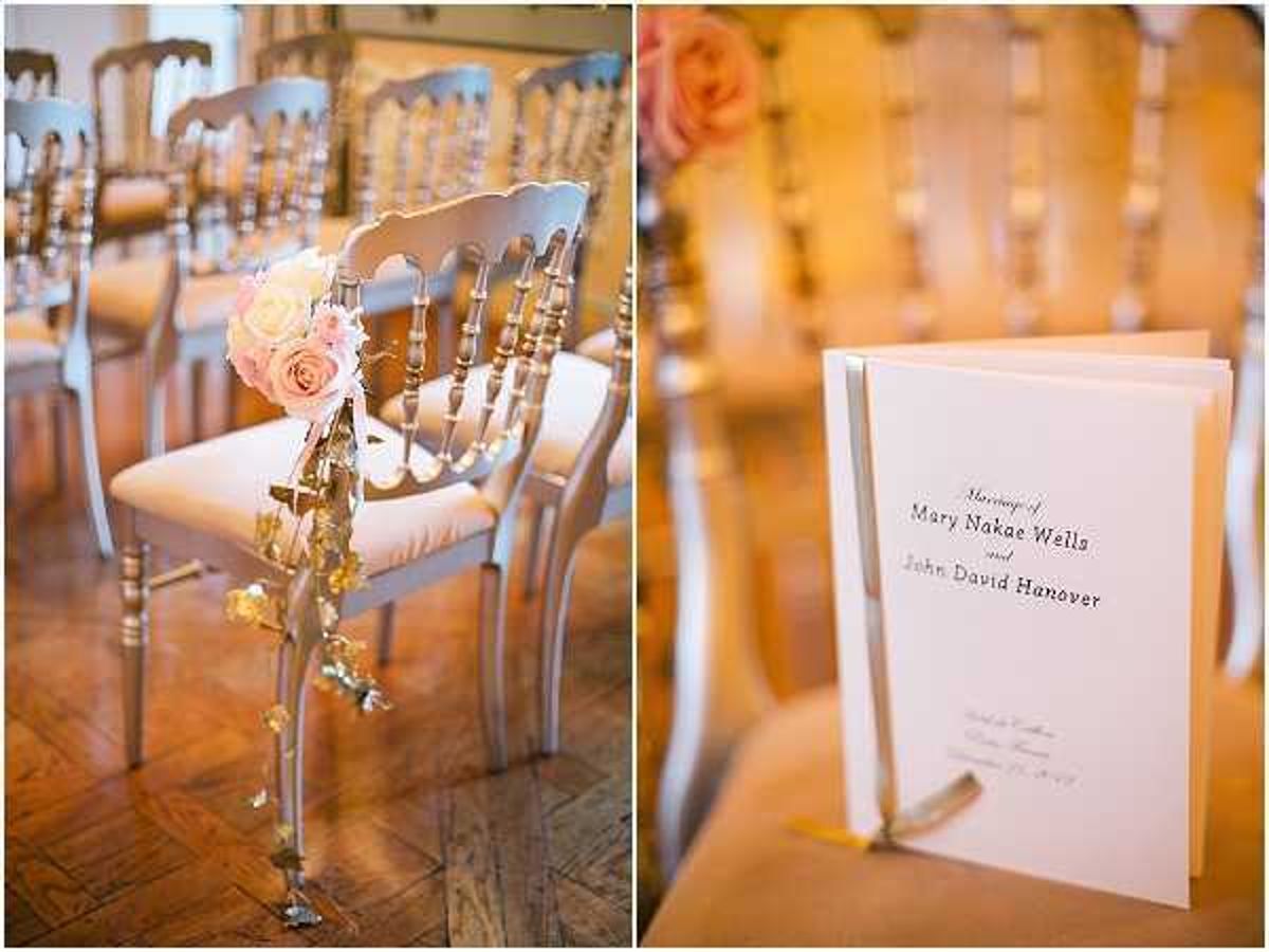 A two-panel detail shot from an indoor wedding ceremony setup. The left panel shows silver Chiavari chairs arranged in rows, with the aisle chair decorated by a small floral cluster of blush and ivory roses tied with gold ribbon and trailing gold leaf garland. The right panel shows a white folded ceremony program placed on a chair seat, printed with the names 'Mary Nakae Wells and John David Hanover' in an elegant serif font. The decor palette combines silver, blush pink, ivory, and gold accents, consistent with a classic, refined ceremony styling.