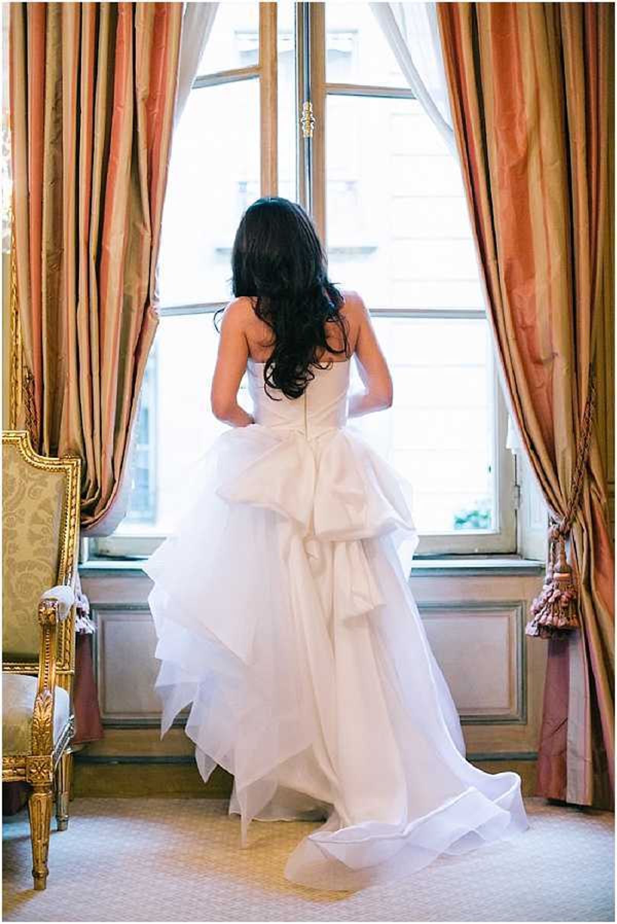 A bridal portrait taken indoors showing the bride from behind as she stands at a tall window looking out. She is wearing a strapless white ball gown with a structured, layered skirt featuring large satin bow detailing at the back and a flowing tulle train. Her long dark hair falls in loose waves down her back. The room features floor-length salmon and gold striped silk curtains with tassel tiebacks, ornate gold-framed upholstered chairs, and wood-paneled walls, suggesting a classic Parisian hotel or chateau interior. The composition is a full-length portrait shot with natural backlight coming through the window.