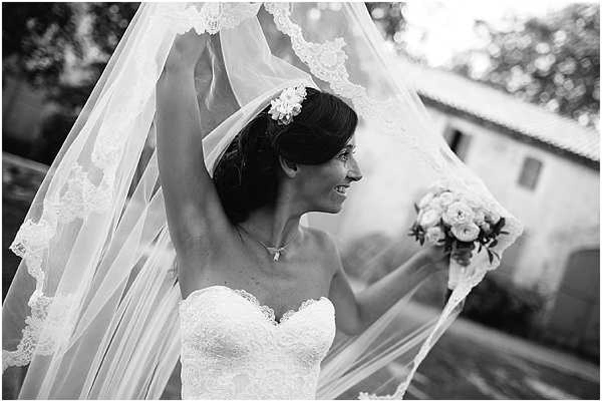A black-and-white bridal portrait taken outdoors, showing the bride from approximately the waist up. She is smiling and holding her long lace-trimmed cathedral veil aloft with one raised arm while holding a round bouquet of roses and smaller blooms in her other hand. She wears a strapless sweetheart-neckline lace gown, a floral hair accessory, and a delicate necklace. Her dark hair is styled in an updo. The veil's wide lace border is prominent in the frame, creating a dramatic flowing effect. The image has strong contrast with bright highlights on the veil against a softly blurred background that includes a light-colored building. This is a close-to-medium portrait composition.
