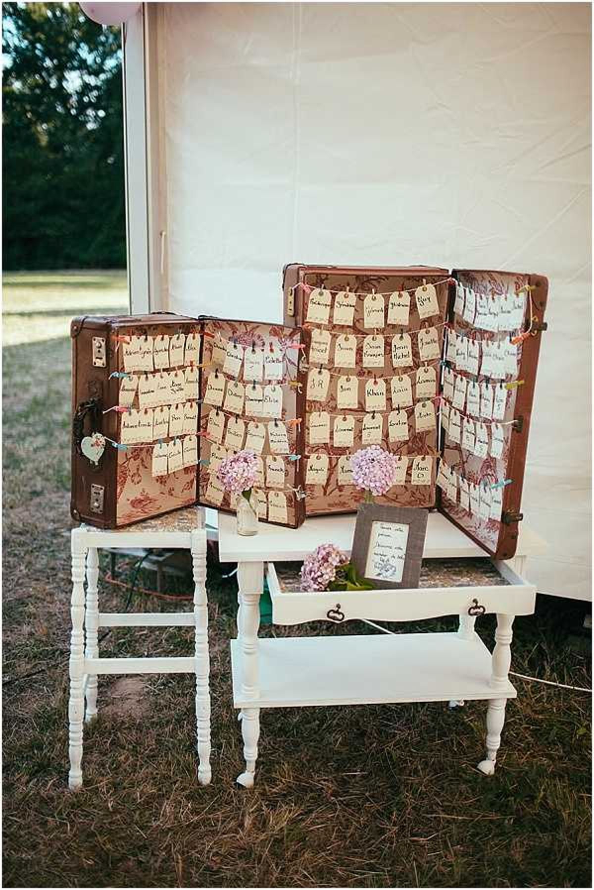 A close-up detail shot of a vintage-styled wedding seating chart display set up outdoors near a white marquee tent. Two antique brown leather trunks are opened and propped up on white painted wooden side tables, with their floral-patterned interiors used as backdrops for rows of kraft paper luggage tags bearing handwritten guest names, strung along colorful twine. Small vases of pink hydrangeas and a handwritten framed note are placed on the table surface between the trunks. The overall styling is rustic-vintage with a travel theme, incorporating warm tones of brown leather, cream paper tags, and soft pink floral accents.