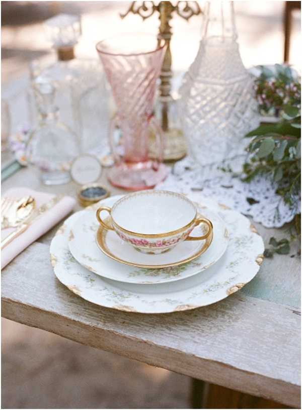 Close-up detail shot of a vintage-style wedding table setting on a whitewashed wooden table. The place setting features stacked china plates with delicate floral and gold trim detailing, topped with a gold-handled teacup and saucer in a matching pattern. A white crocheted doily runner is visible beneath glassware in the background, including a pink twisted-glass vase, a cut-crystal decanter, and a white pressed-glass vessel. A blush pink linen napkin with gold cutlery is placed to the left, along with a small circular place card or wax seal. Greenery, likely eucalyptus, is partially visible at the right edge. The overall styling is romantic vintage with a palette of blush pink, ivory, and gold.